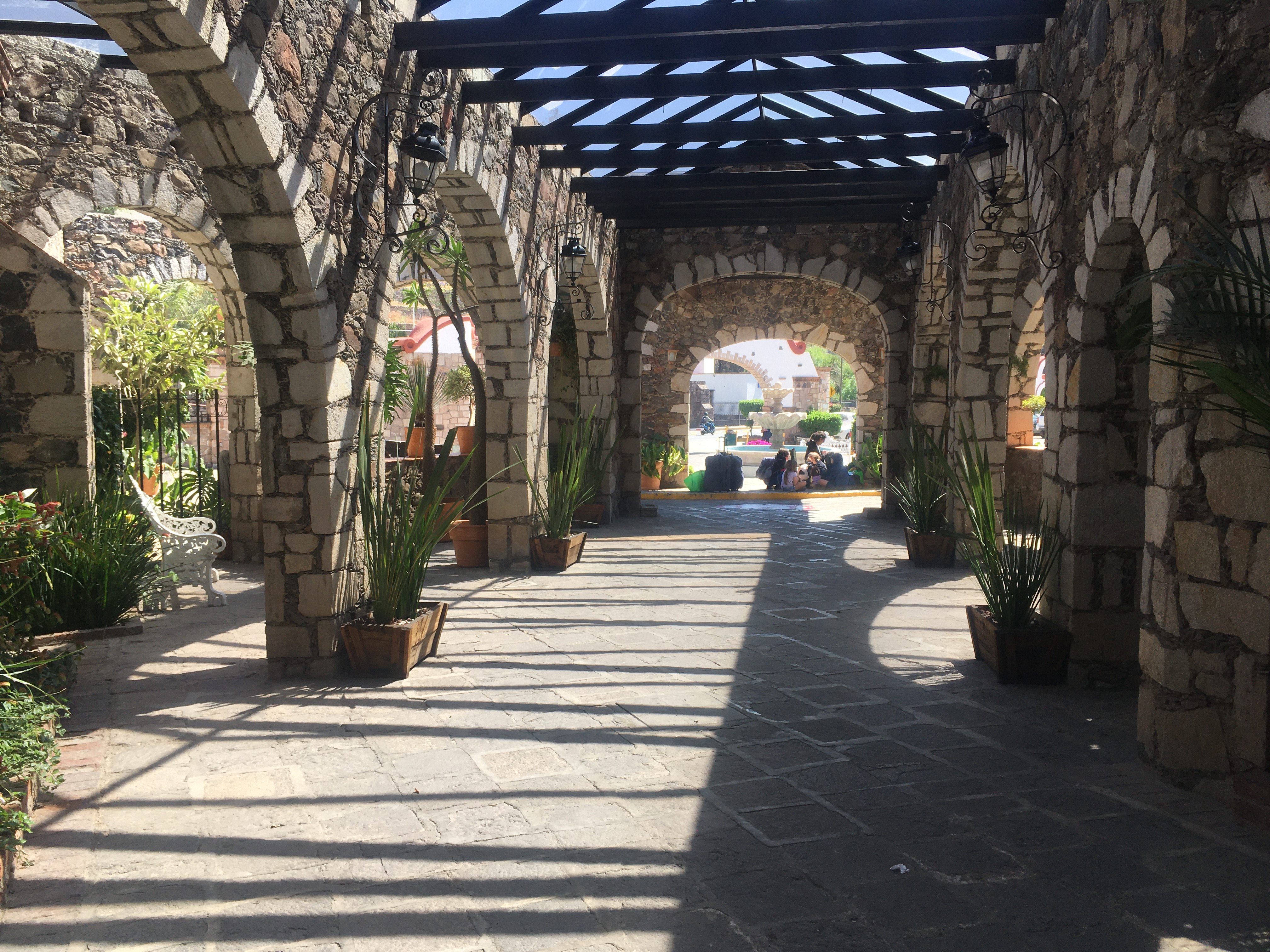 A walkway with stone arches along both sides. There are potted plants, a white wrought-iron bench, and a glimpse of a garden beyond the fence on one side. At the end of the corridor there are two arches, one beyond the other, and a pile of bags and kids sitting at the curb.