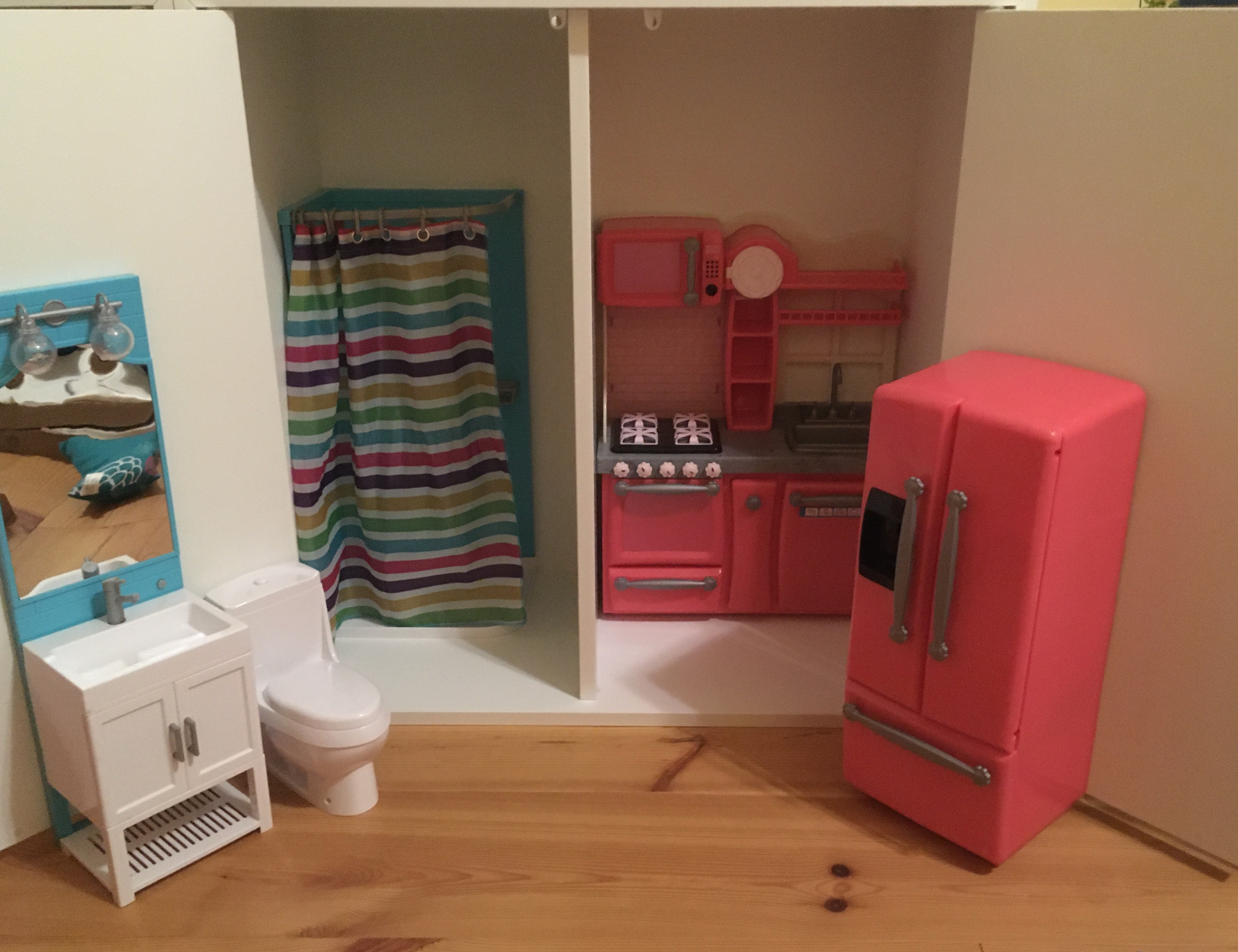 A square cupboard, open, with a doll kitchen in one half and a doll bathroom in the other. The two are separated by a vertical shelf.