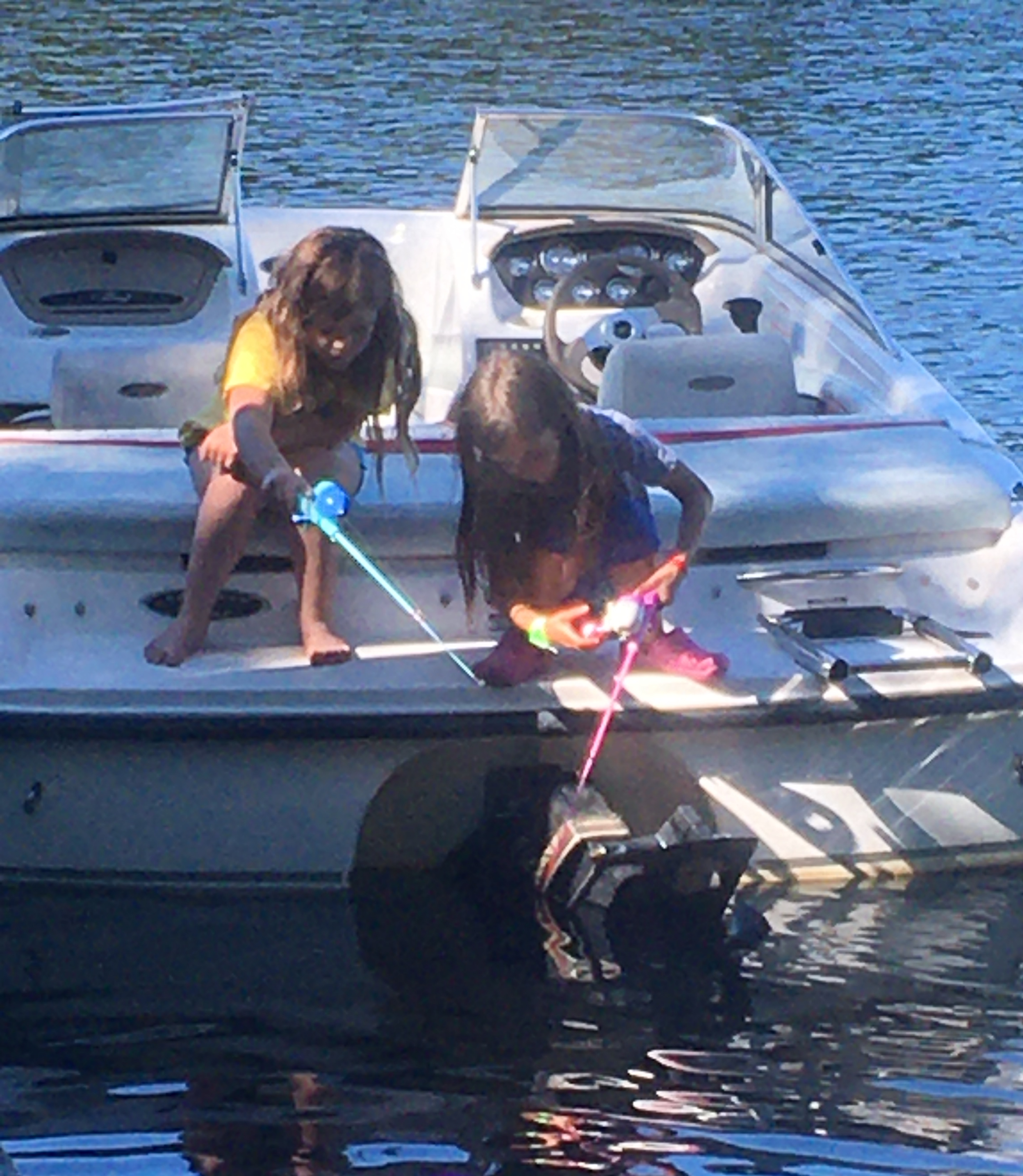 E and her friend sitting on the back of the motorboat with plastic fishing rods.