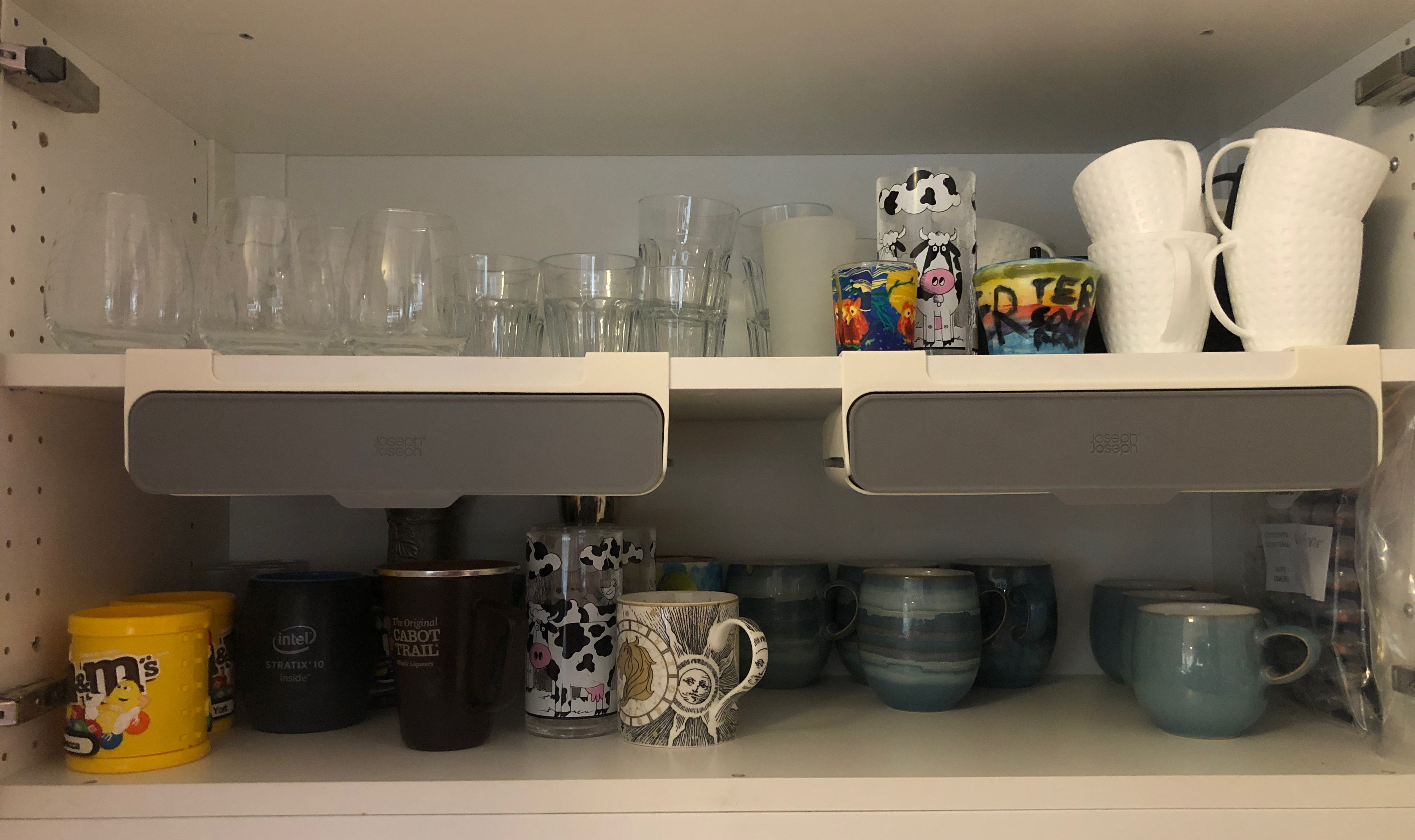 Interior of a cabinet with two shelves holding glasses and mugs. On the underside of the upper shelf are two grey-and-white rectangular containers.